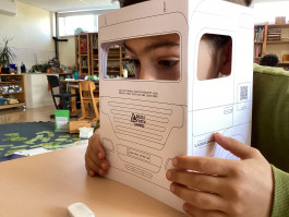 Ein Schüler schaut durch eine Pappe, die wie ein LKW-Fahrerhaus aussieht und den Toten Winkel simuliert